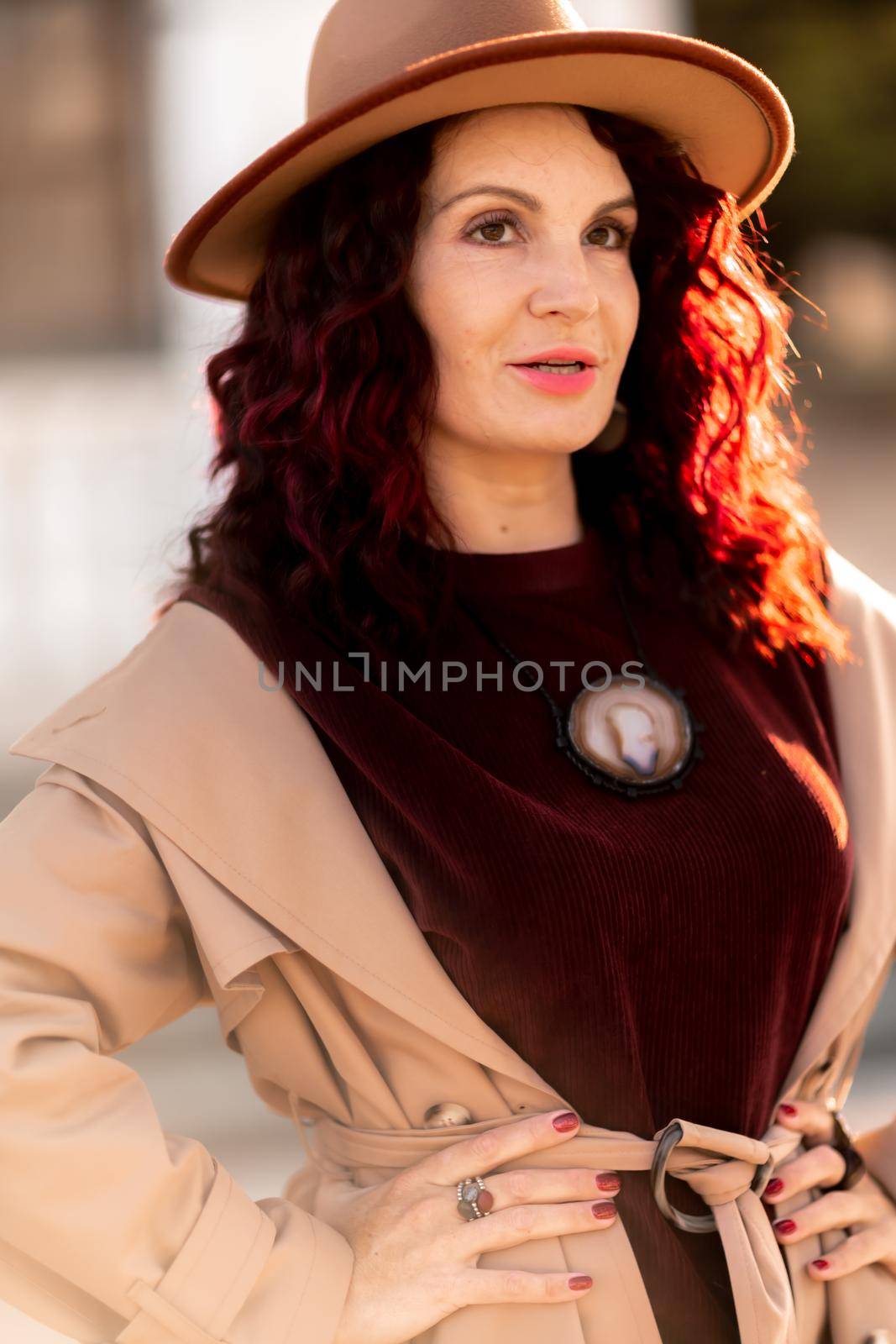 Outdoor fashion portrait of young elegant fashionable brunette woman, model in stylish hat, choker and light raincoat posing at sunset in European city