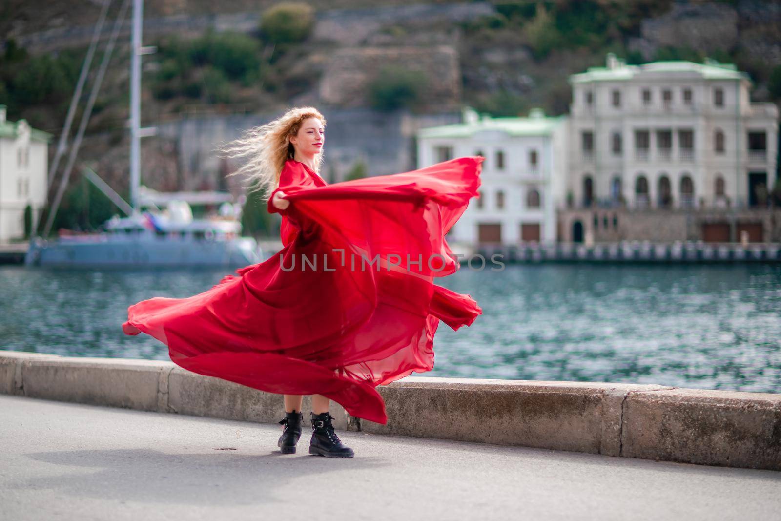 A woman in a red dress, a fashion model with long silk wings in a flowing dress, flying fabric on the embankment