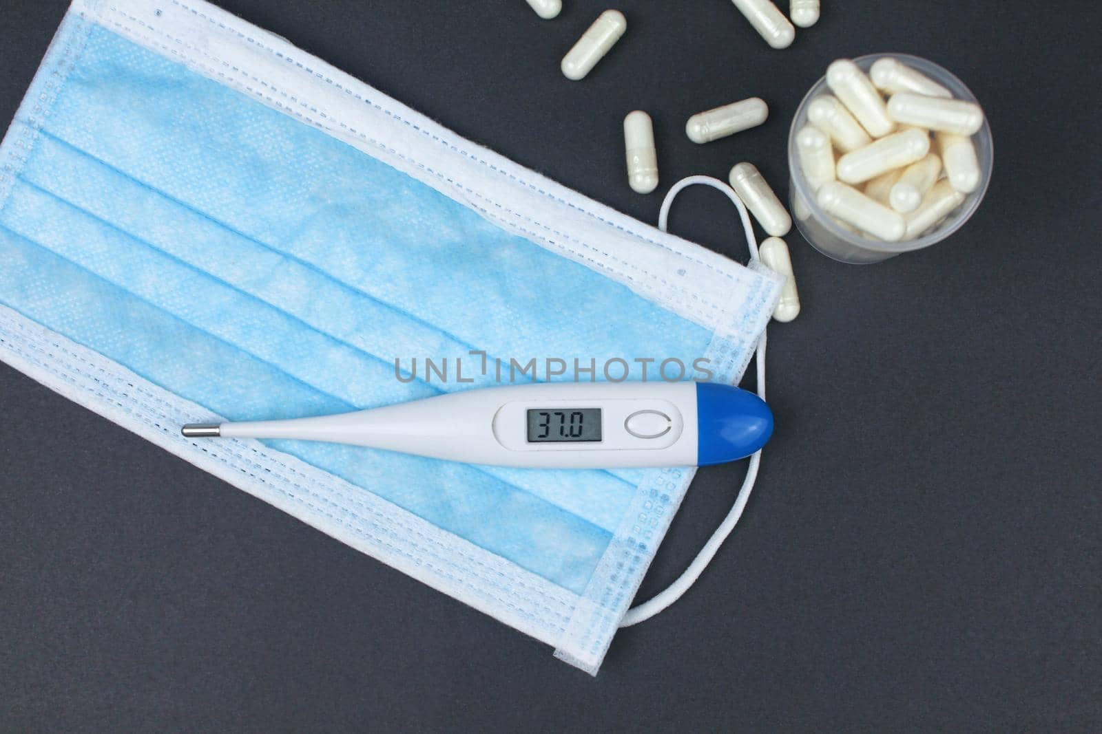White electronic thermometer with packs of tablets and blue medical face mask on black background.