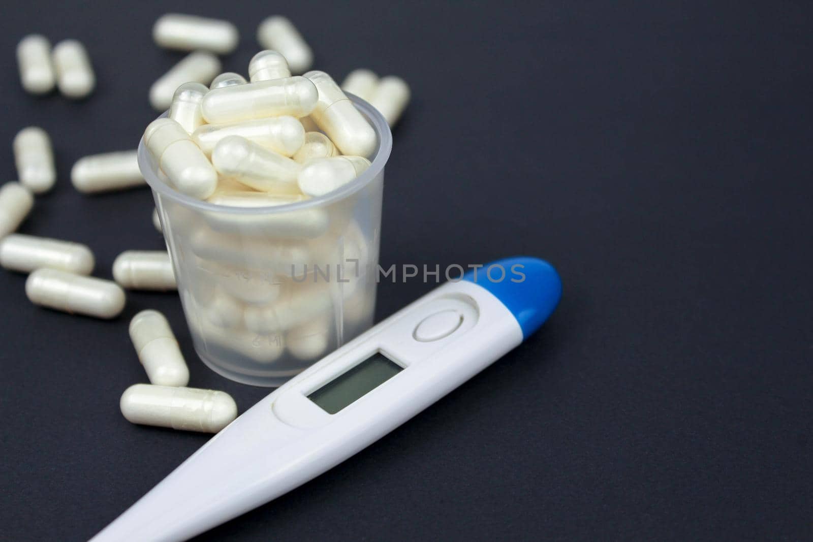White electronic thermometer and pills in a measuring cup on a black background. Text for space. Selective focus. by JuliaDorian