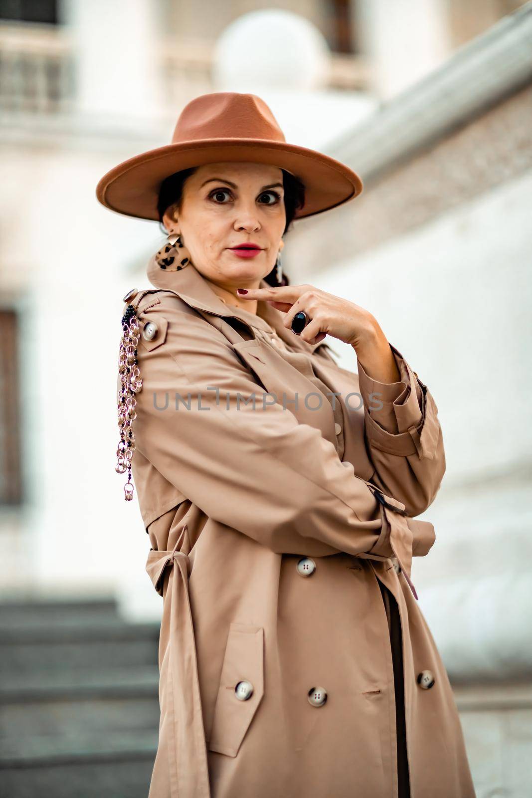 Outdoor fashion portrait of young elegant fashionable brunette woman, model in stylish hat, choker and light raincoat posing at sunset in European city