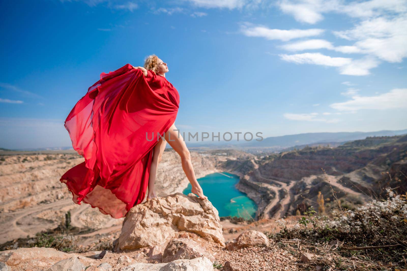 Side view of a beautiful sensual woman in a red long dress posing on a rock high above the lake in the afternoon. Against the background of the blue sky and the lake in the form of a heart by Matiunina