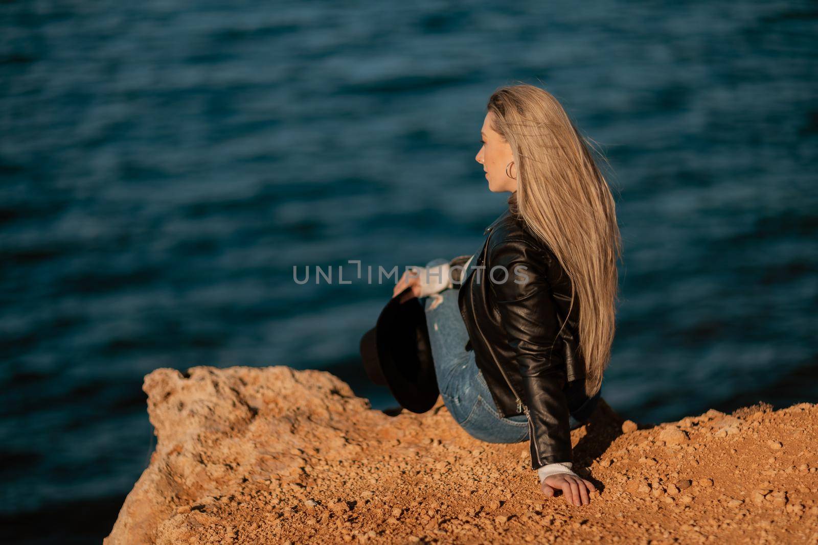 A blonde girl in a stylish black leather jacket is sitting with her back to the seashore. by Matiunina