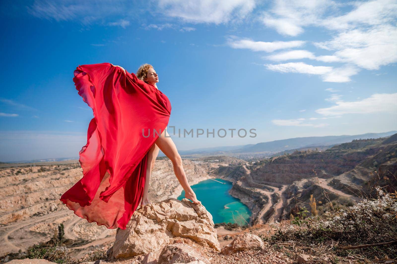Side view of a beautiful sensual woman in a red long dress posing on a rock high above the lake in the afternoon. Against the background of the blue sky and the lake in the form of a heart by Matiunina