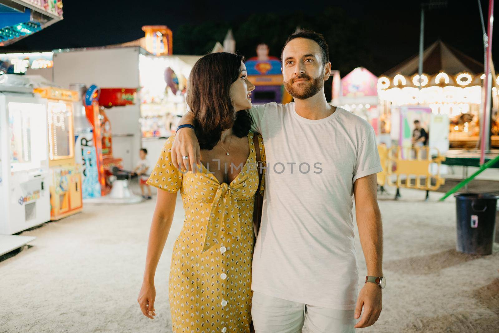 A girl in a yellow dress with a plunging neckline and her boyfriend with a beard are hugging each other and walking between amusement rides. A couple of lovers on a date at the fair in Valencia.