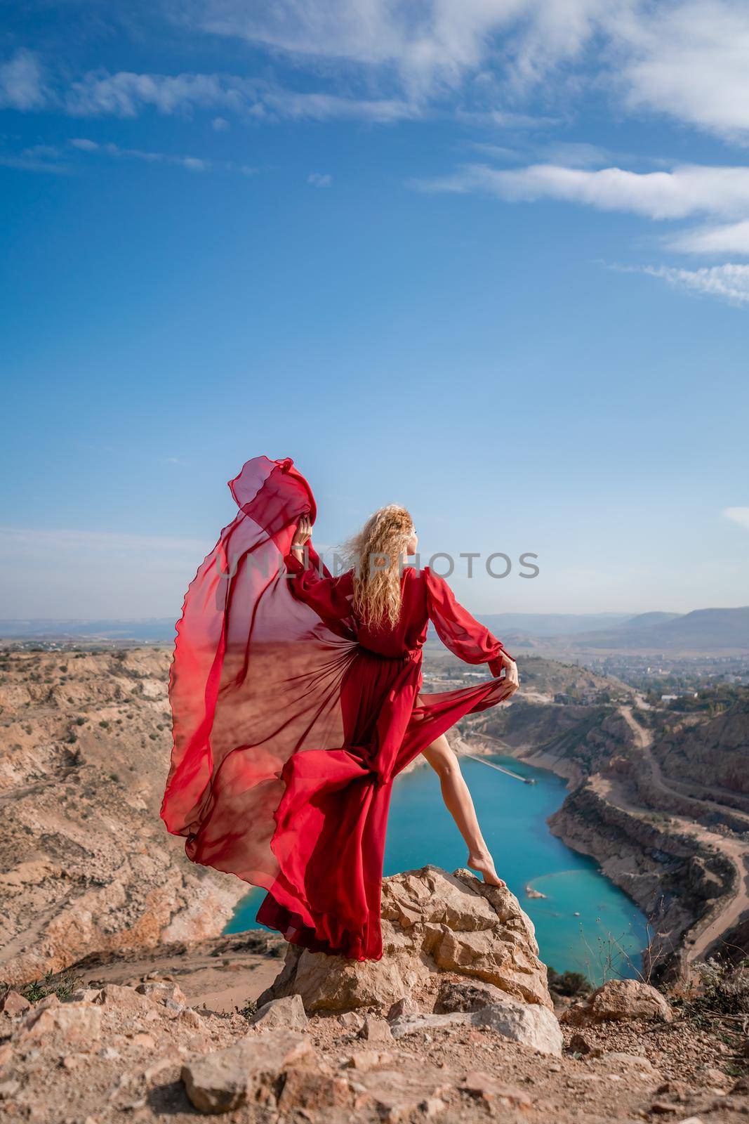 Rear view of a beautiful sensual woman in a red long dress posing on a rock high above the lake in the afternoon. Against the background of the blue sky and the lake in the shape of a heart by Matiunina