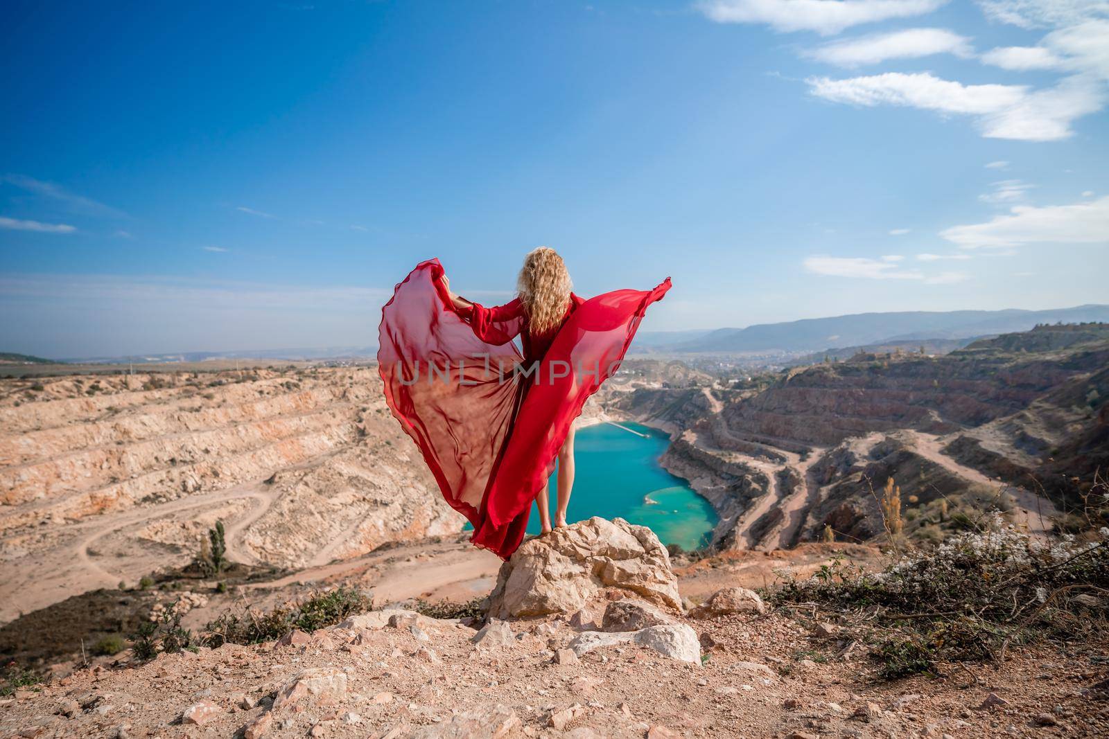 Side view of a beautiful sensual woman in a red long dress posing on a rock high above the lake in the afternoon. Against the background of the blue sky and the lake in the form of a heart.