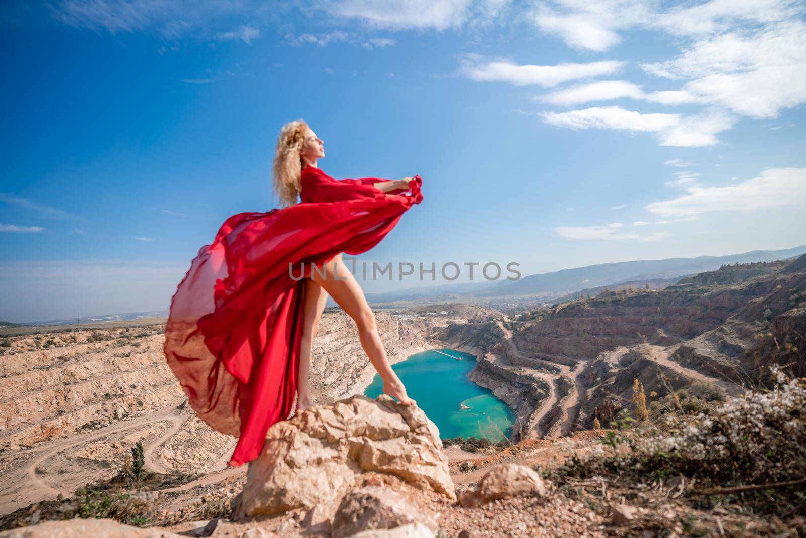 Side view of a beautiful sensual woman in a red long dress posing on a rock high above the lake in the afternoon. Against the background of the blue sky and the lake in the form of a heart by Matiunina