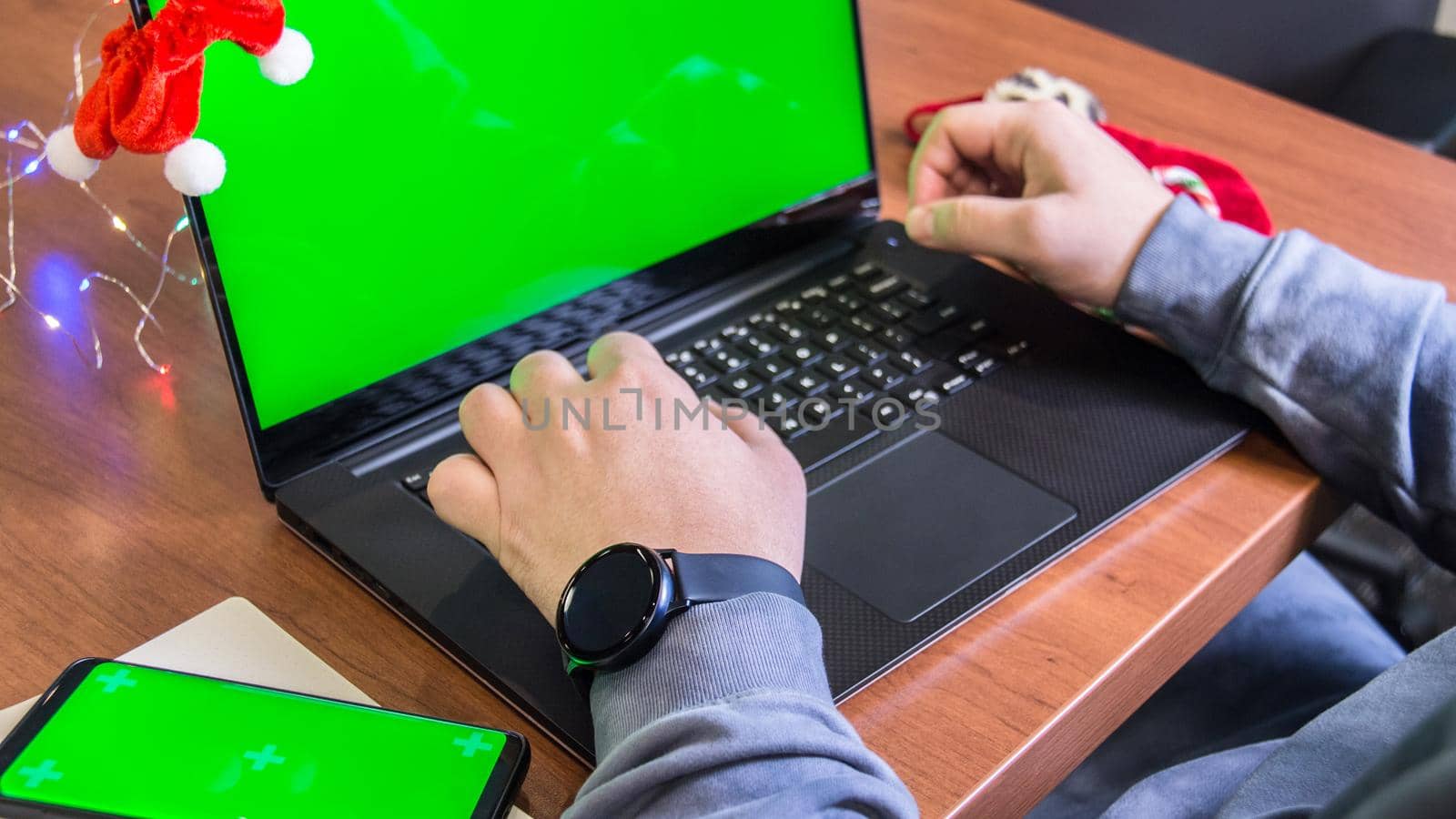 Closeup man hands typing on laptop keyboard with empty green screen, smartphone mockup and Christmas decoration on background. Holiday concept. Office. Mock-up.