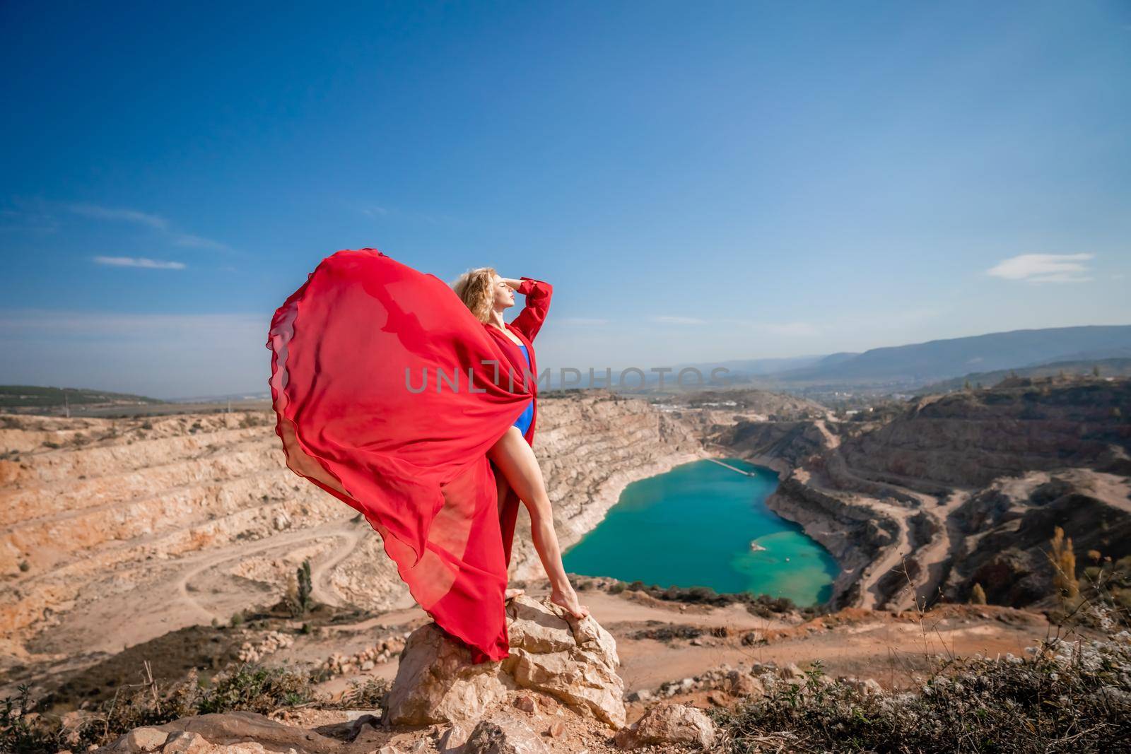 Side view of a beautiful sensual woman in a red long dress posing on a rock high above the lake in the afternoon. Against the background of the blue sky and the lake in the form of a heart by Matiunina