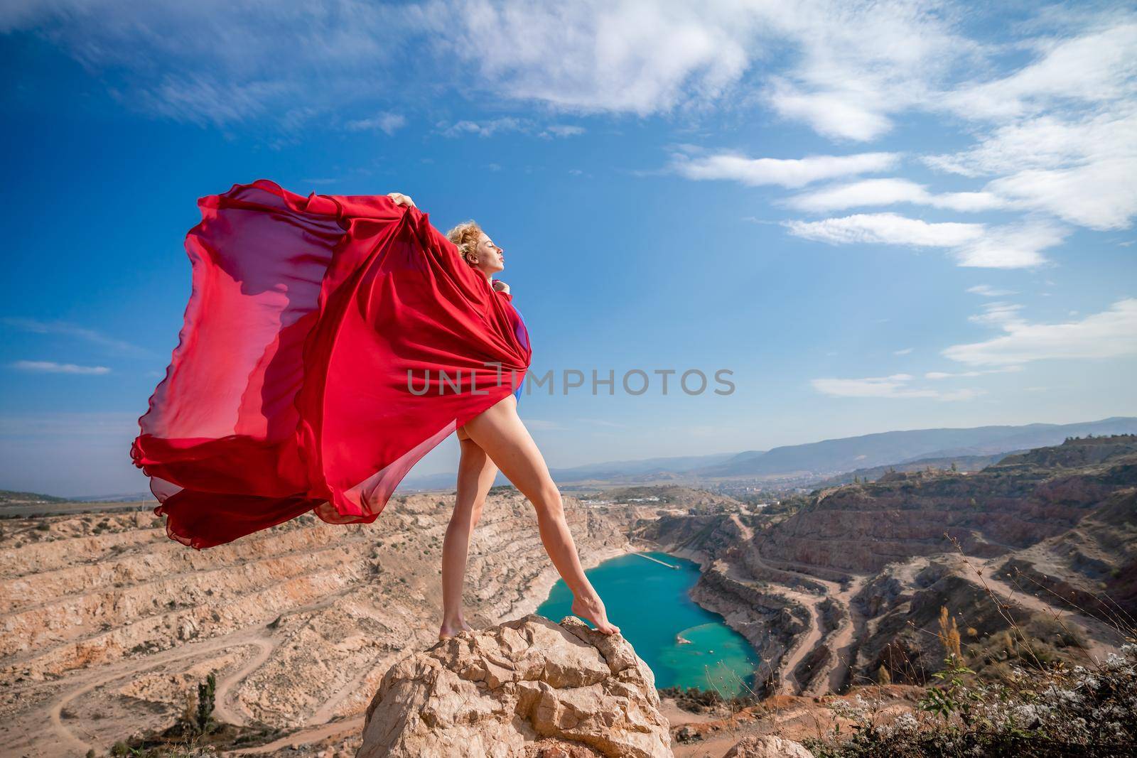 Side view of a beautiful sensual woman in a red long dress posing on a rock high above the lake in the afternoon. Against the background of the blue sky and the lake in the form of a heart.