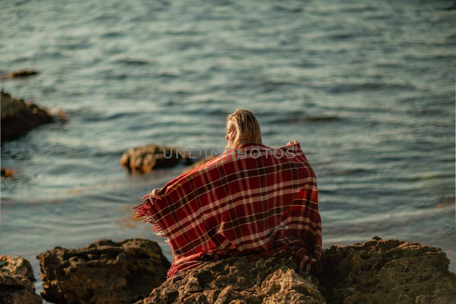 Attractive blonde Caucasian woman enjoying time on the beach at sunset, sitting in a blanket and looking to the side, with the sunset sky and sea in the background. Beach vacation. by Matiunina