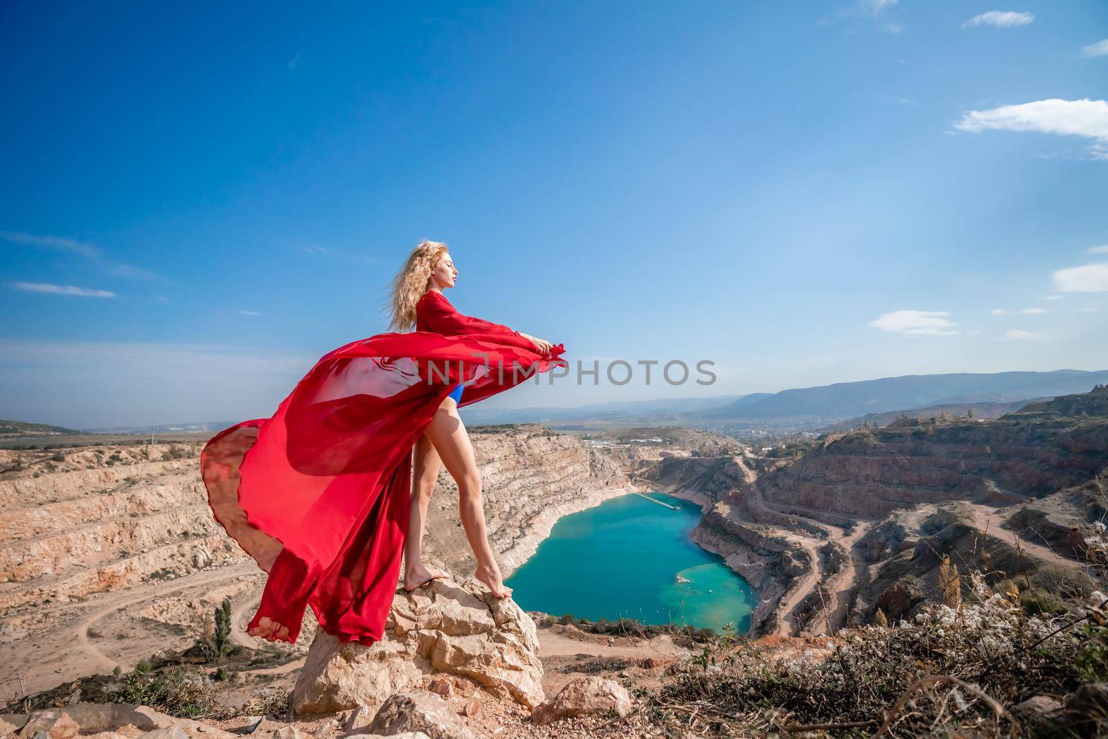 Side view of a beautiful sensual woman in a red long dress posing on a rock high above the lake in the afternoon. Against the background of the blue sky and the lake in the form of a heart.