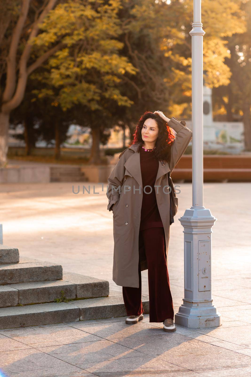 Outdoor fashion portrait of young elegant fashionable brunette woman, model in stylish hat, choker and light raincoat posing at sunset in European city