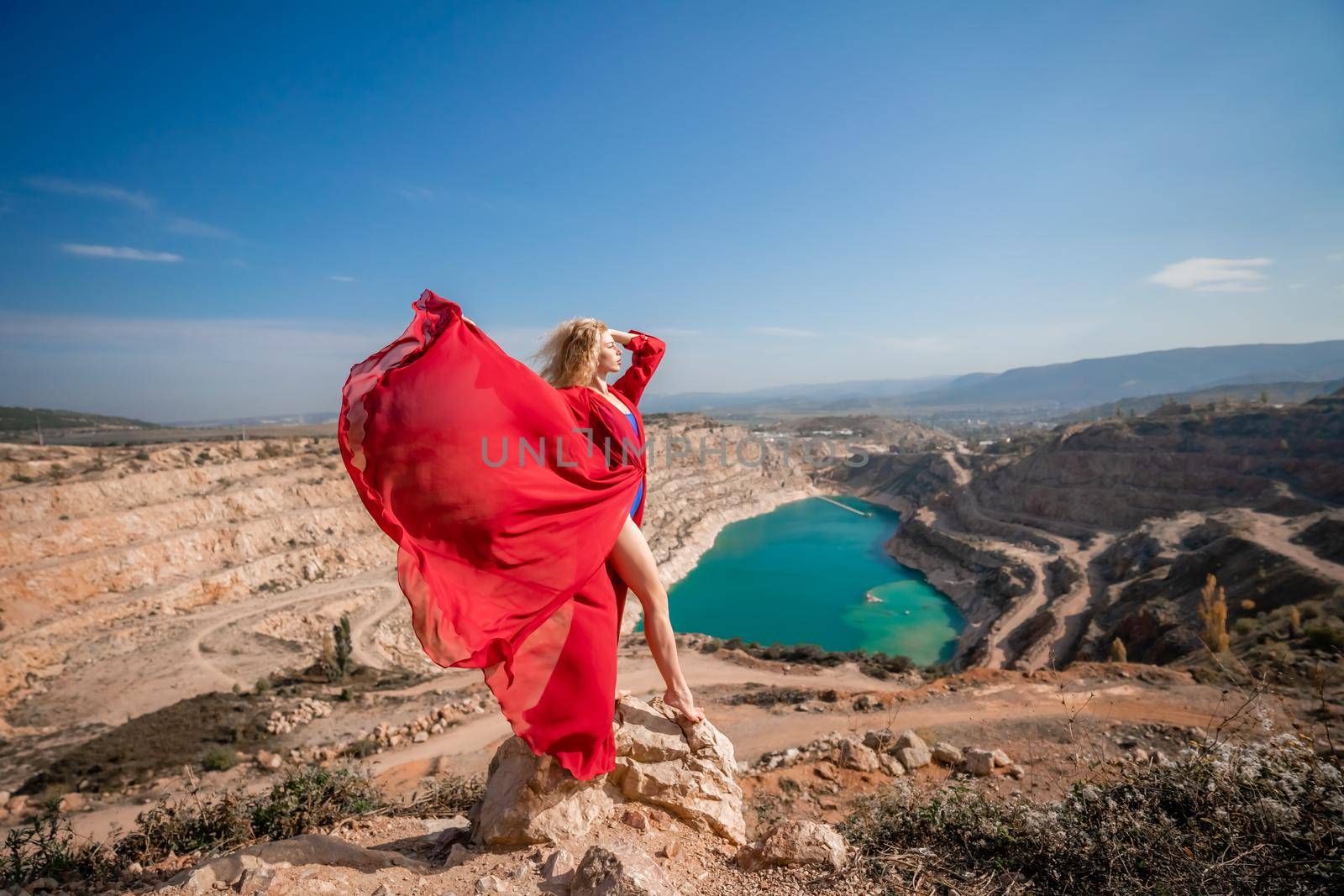 Side view of a beautiful sensual woman in a red long dress posing on a rock high above the lake in the afternoon. Against the background of the blue sky and the lake in the form of a heart by Matiunina