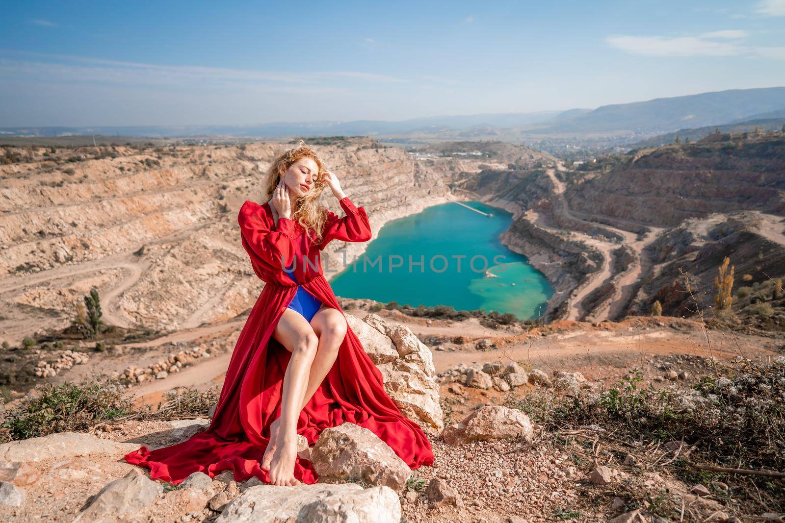 A beautiful girl in a red long dress, Sits on a rock high above the lake in the afternoon. Against the background of the blue sky and the lake in the shape of a heart