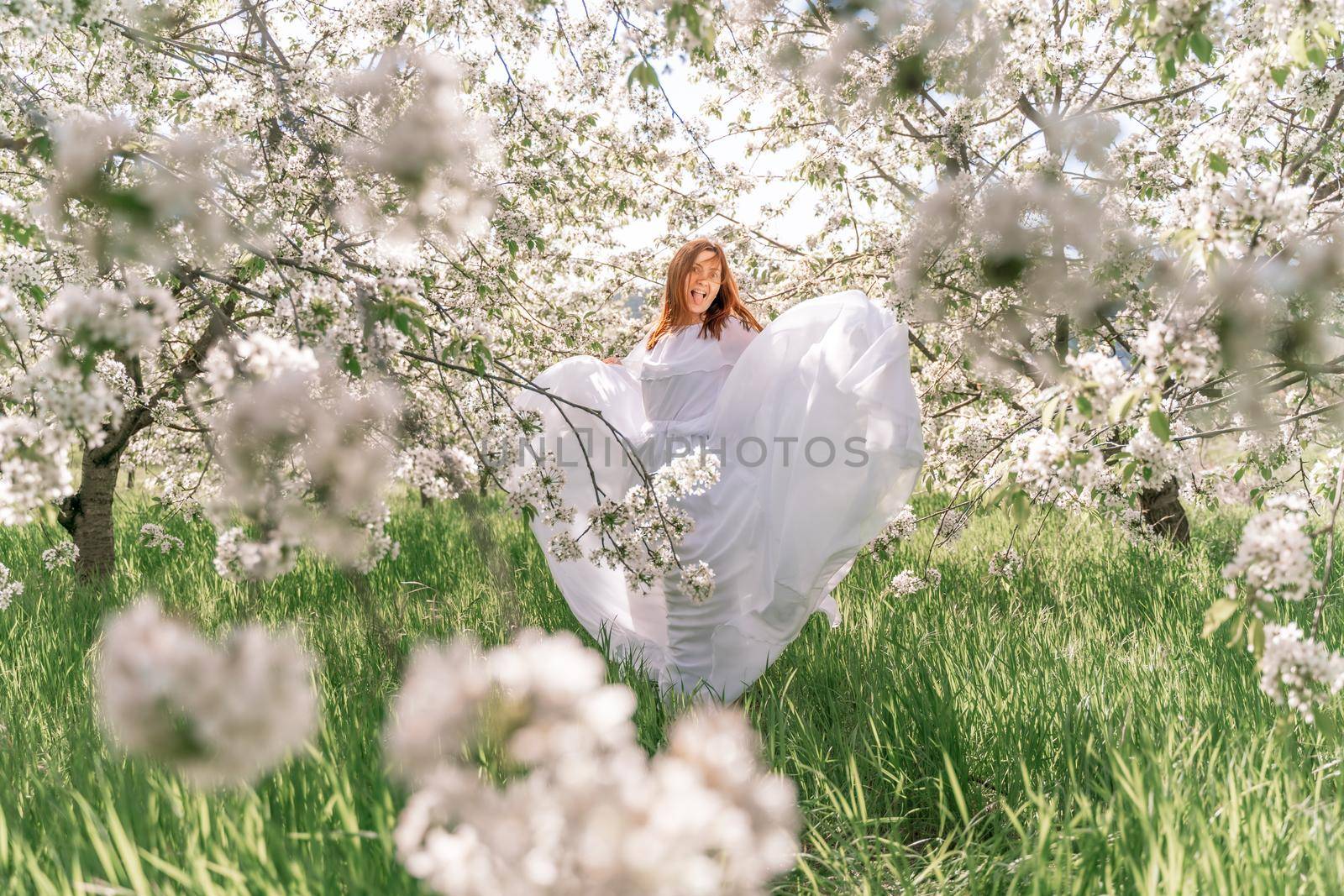 Fantasy woman in long white elegant fashion long dress walks in green spring blossom cherry garden. Happy cheerful girl princess bride. Skirt fabric flies flowing waving in wind motion. by Matiunina