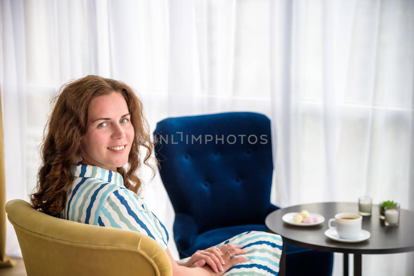 Young woman at home sitting on modern chair in front of window relaxing in her living room and drinking coffee or tea with sweets or desert. Relax before new business day.