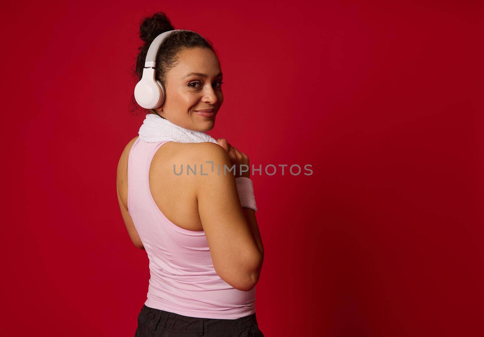 Charming mature African American active woman in sportswear, wireless headphones, smiles, looking over shoulders at camera, relaxes after workout, isolated on red wall background