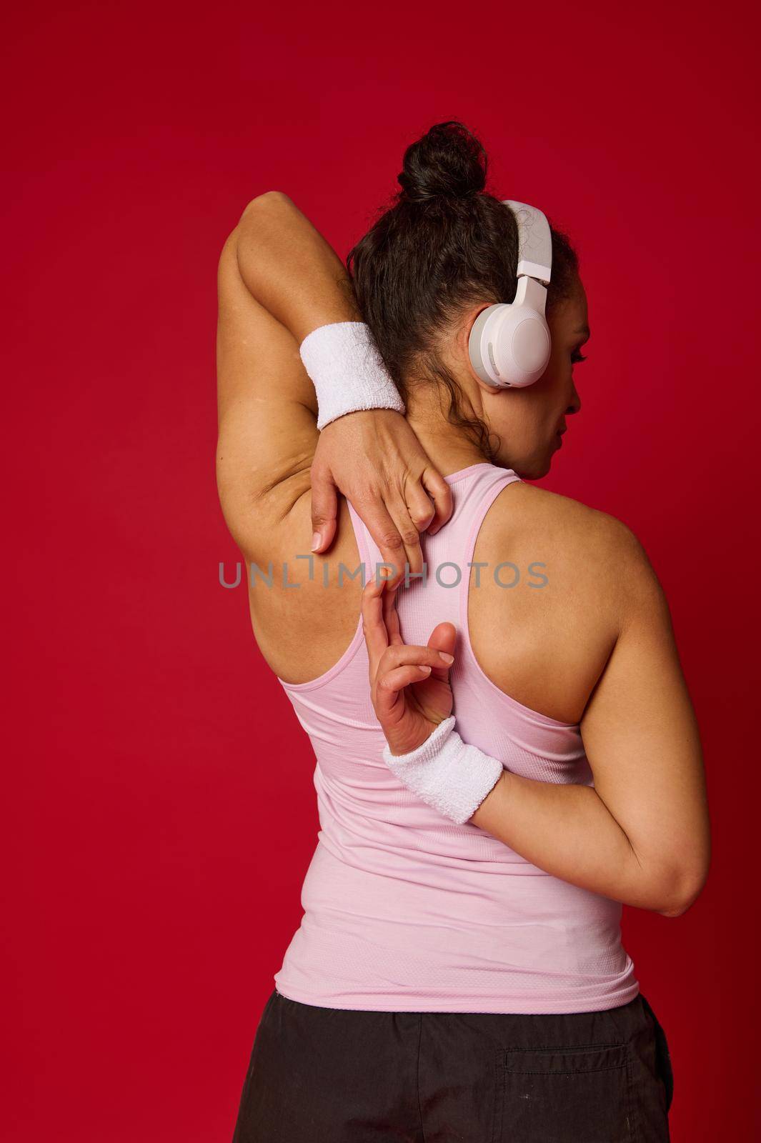 Portrait of a sportswoman in activewear and wireless headphone doing stretching exercises on arms standing back to the camera against red background by artgf