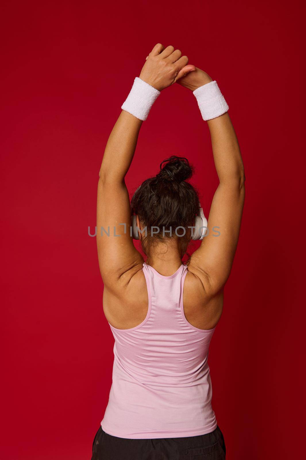 Portrait of female athlete in activewear and wireless headphone doing stretching exercises on arms standing back to the camera against red background by artgf