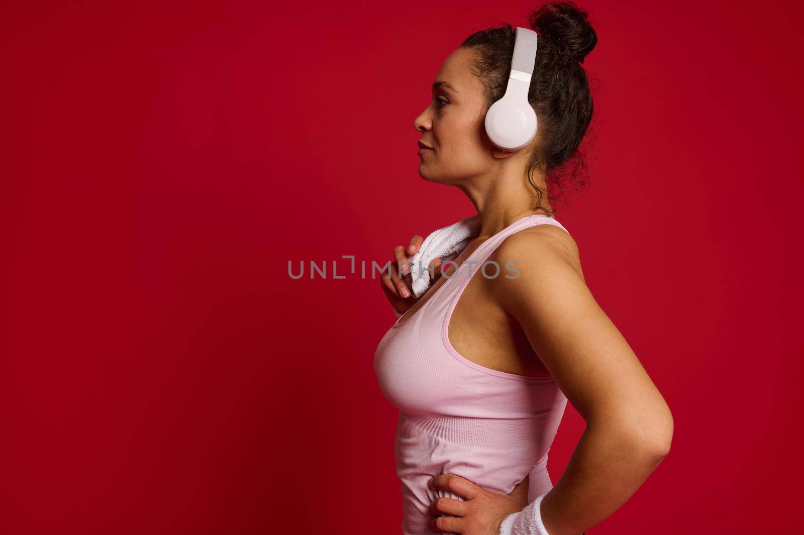 Side portrait of an attractive Latin American sporty woman with white wireless headphones and terry towel on her shoulders, isolated over red colored background with copy ad space