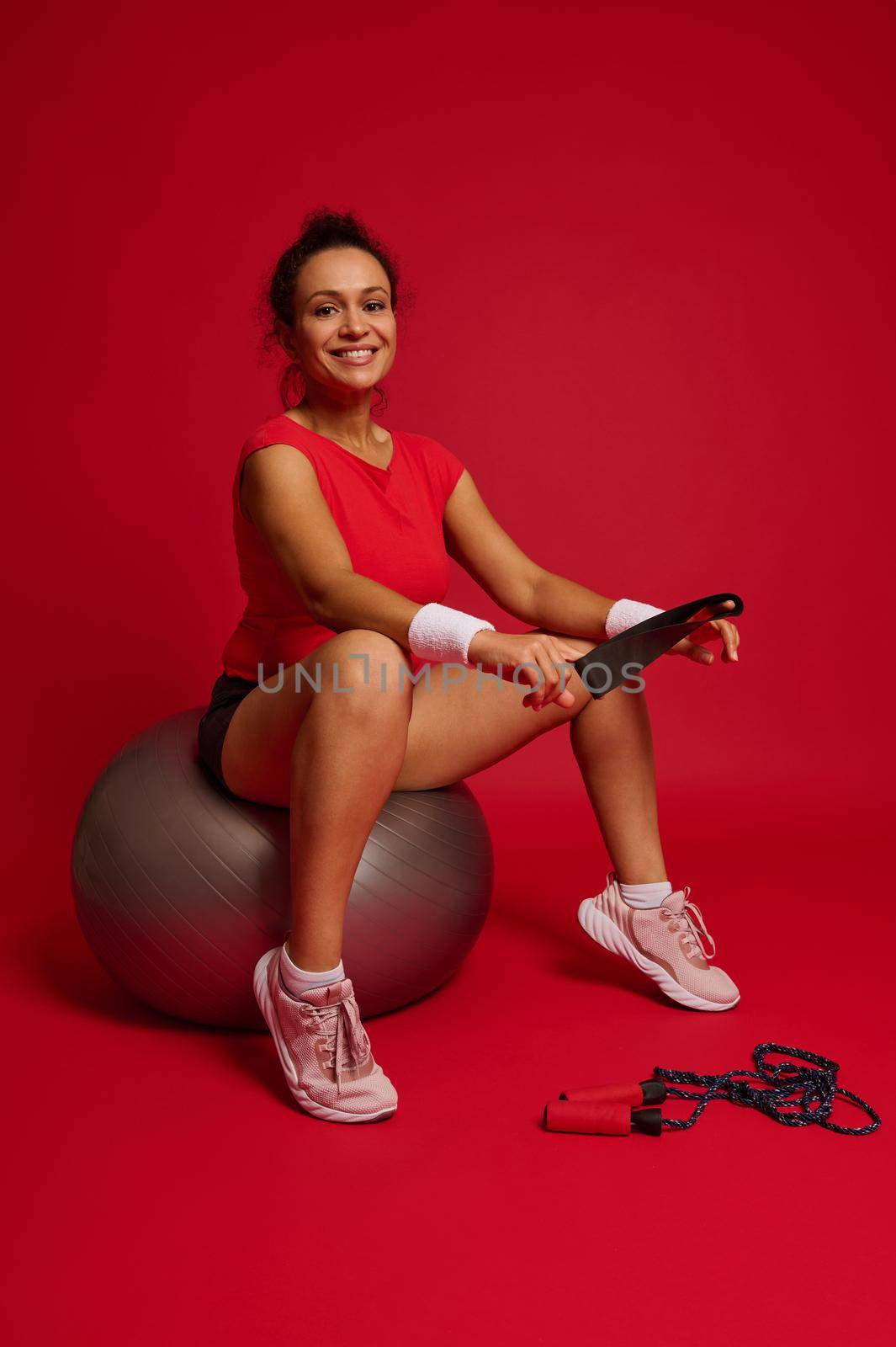 Charming stunning Hispanic sportswoman holding elastic fitness band and sitting on fitness ball after exercising isolated on red background. Concept of slimness, healthy lifestyle and workout. by artgf