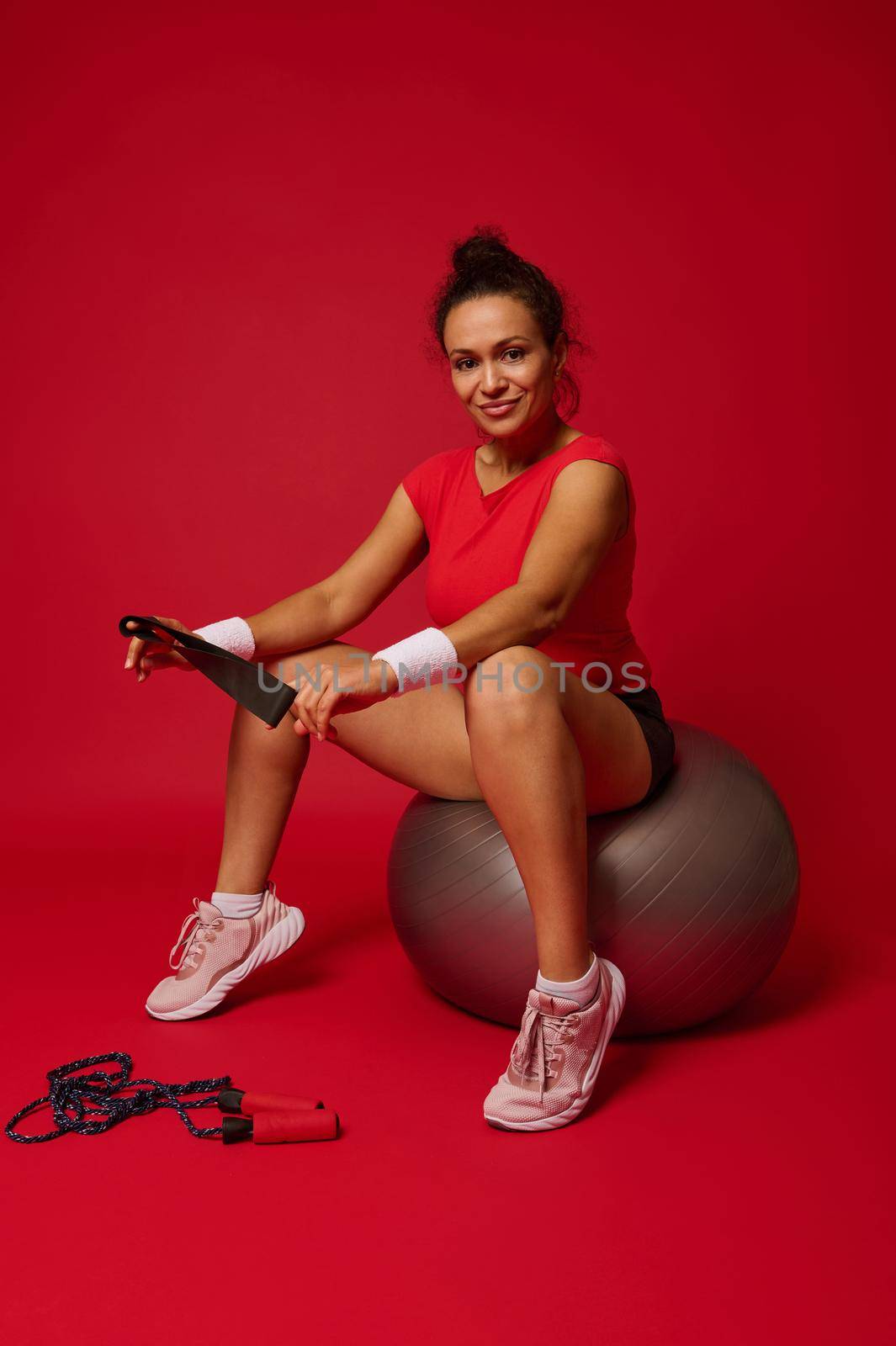 Hispanic beautiful smiling athletic woman holding elastic fitness band and sitting on fitness ball after exercising isolated on red background. Concept of slimness, healthy lifestyle and workout.