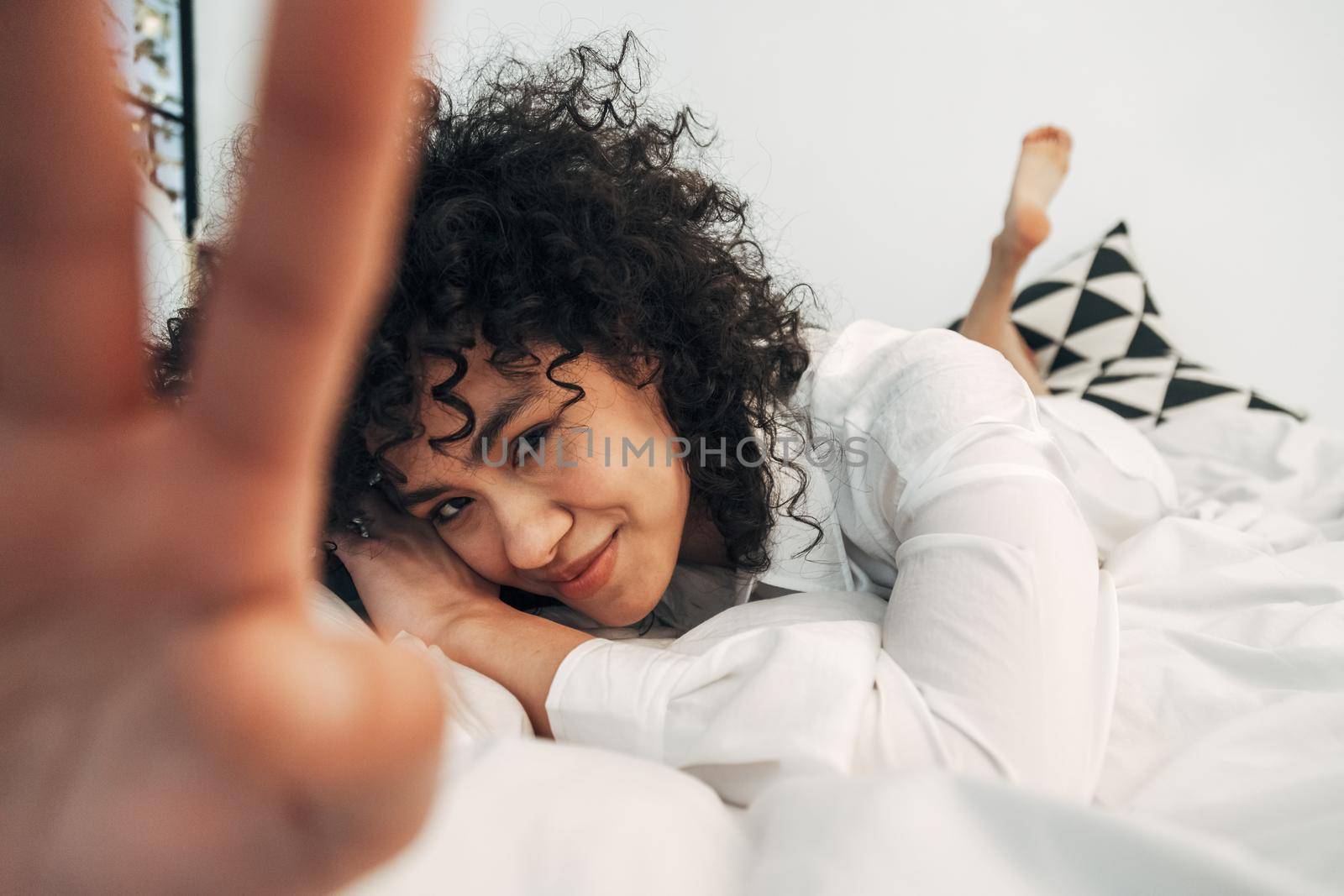 Shy young mixed race woman looking at camera lying down on the bed covering lens with hand. Selective focus on face. by Hoverstock