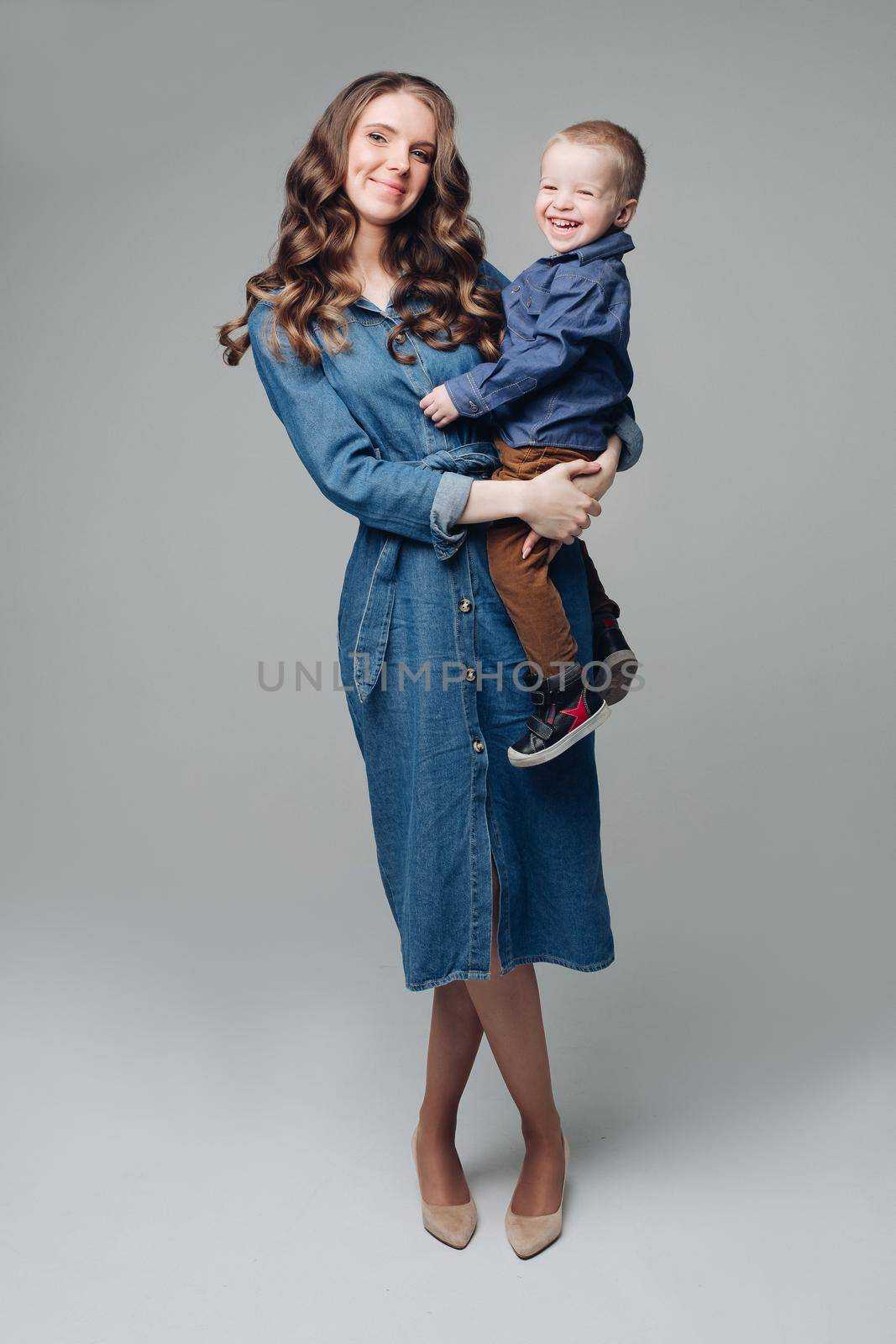 Full length studio portrait of gorgeous elegant brunette mom in denim dress with lovely laughing son on her hands. They smiling at camera.