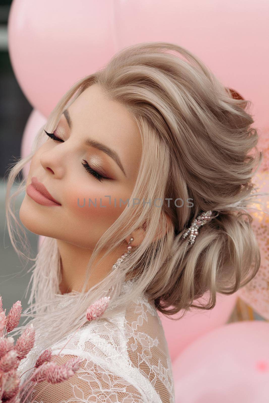 Fashion portrait of stunning blonde girl with hairstyle and professional make up in white transparent blouse holding bunch of pink colored flowers. Bunch of pink air balloons in background.