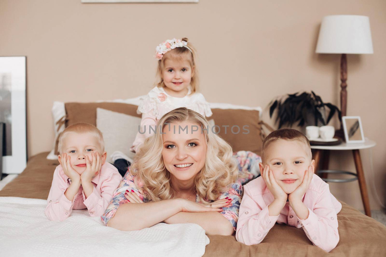 Front view of attractive mother posing with three cute kids at home. Young woman with sweet daughter and little boys looking at camera and smiling. Concept of childhood and motherhood.