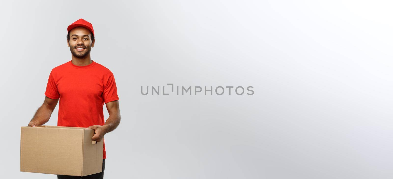 Delivery Concept - Portrait of Happy African American delivery man in red cloth holding a box package. Isolated on Grey studio Background. Copy Space. by Benzoix