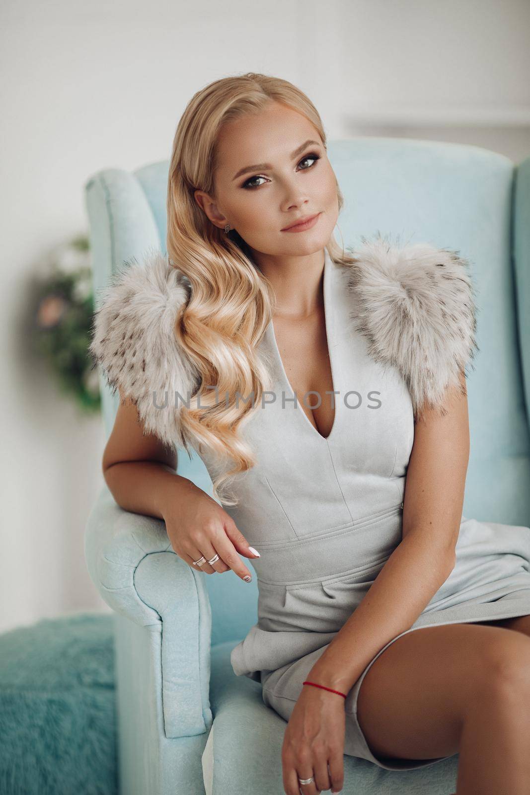 Front view of beautiful blonde in blue short dress and heels sitting on dressing table and posing. Attractive woman with long legs looking down and smiling at home. Concept of beauty and glamour.