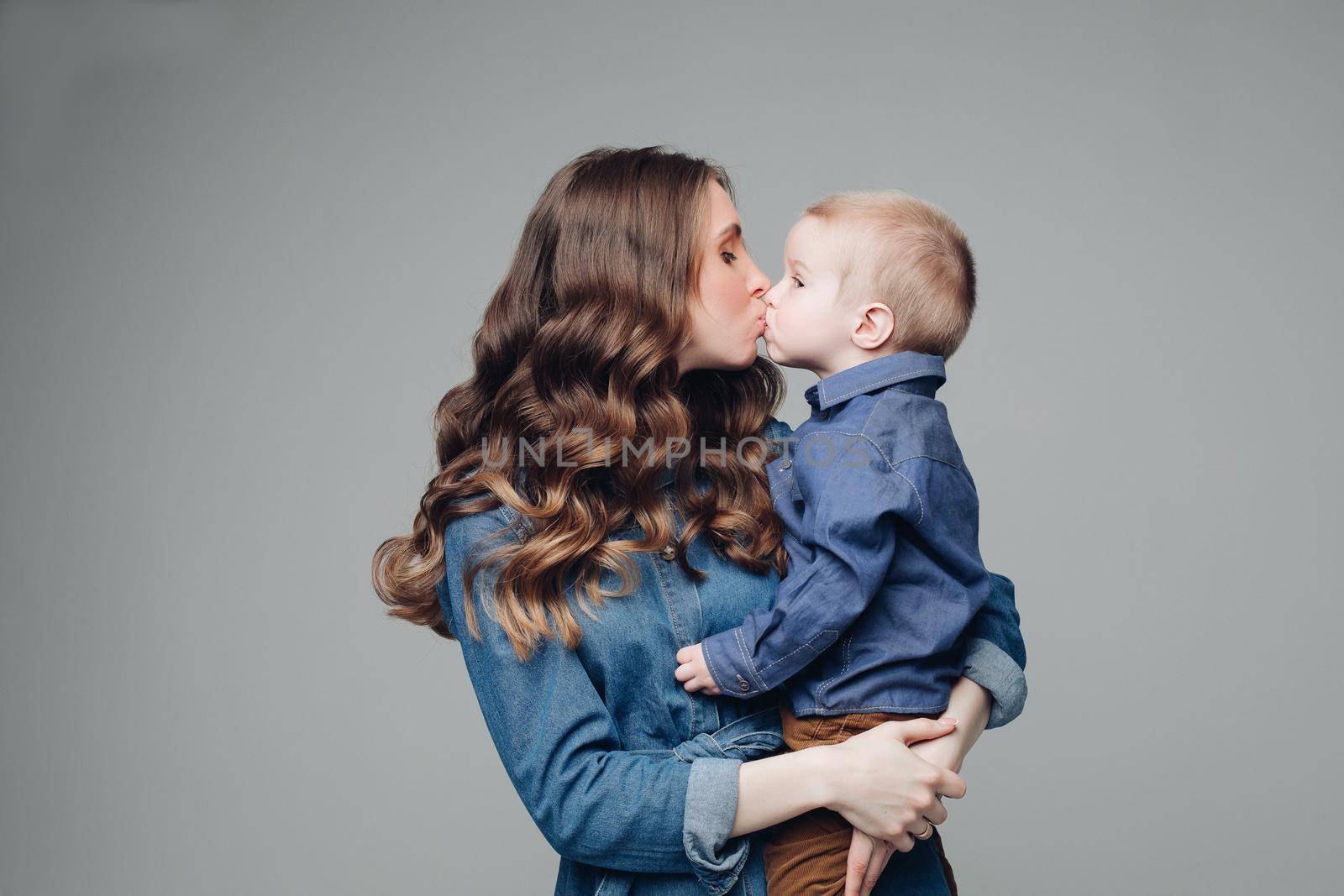 Full length studio portrait of gorgeous elegant brunette mom in denim dress with lovely laughing son on her hands. They smiling at camera.