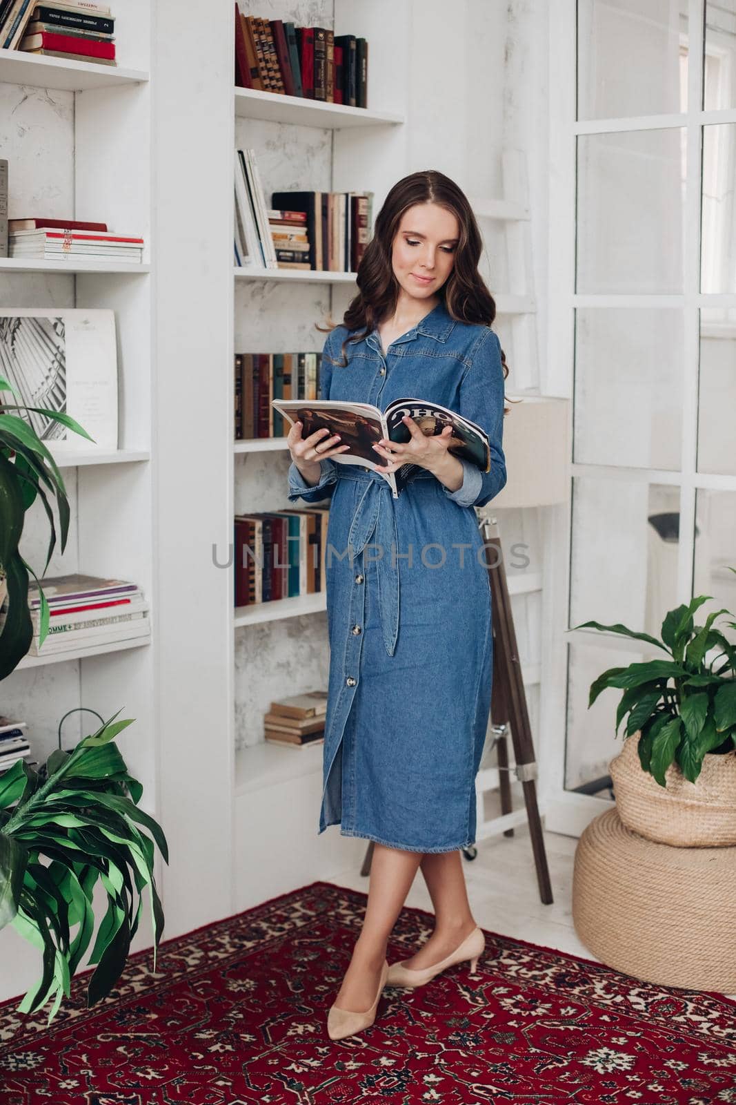 Full length of attractive elegant brunette young woman in stylish casual denim dress and beige heels reading a fashion magazine. She is in living room next to the bookshelf and plants.