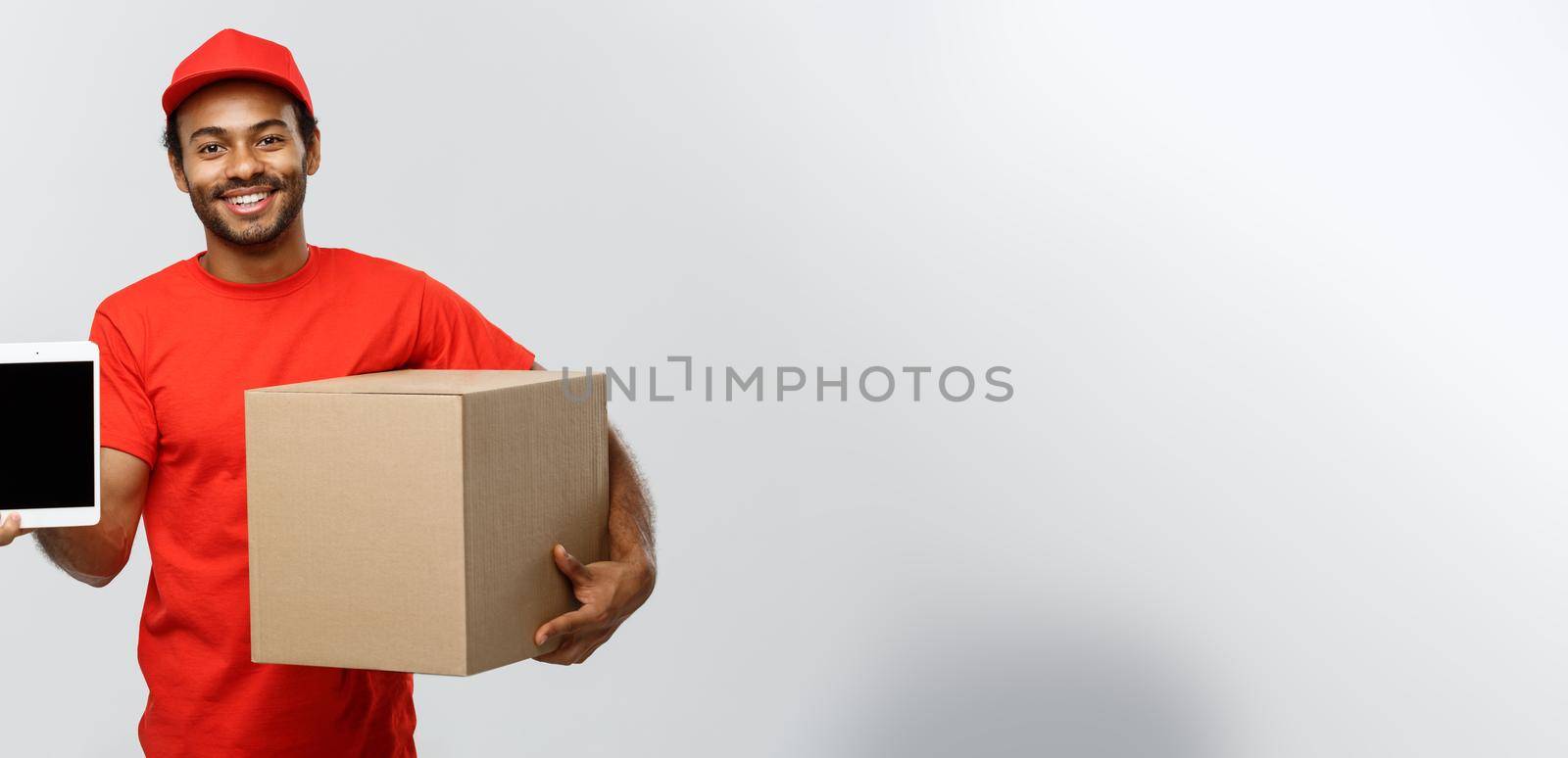 Delivery Concept - Portrait of Handsome African American delivery man or courier with box showing tablet on you to check the order. Isolated on Grey studio Background. Copy Space. by Benzoix