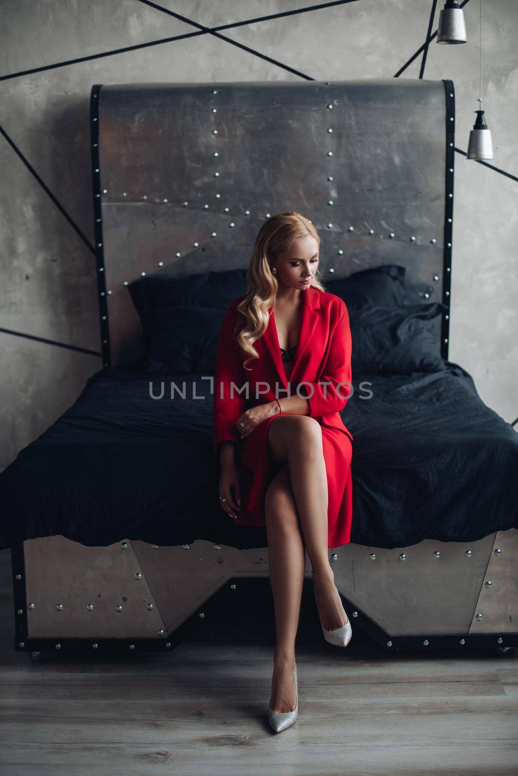 Gorgeous blonde woman sitting on high chair and thoughtfully looking at camera. Young pretty lady in red dress with naked shoulder touching floor by feet. Elegant model holding hand near face posing.