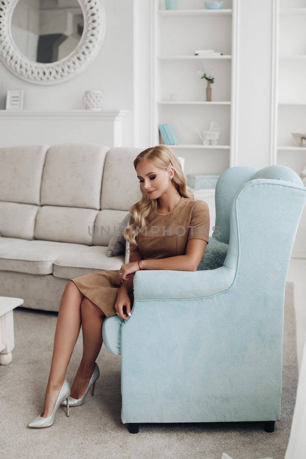 Gorgeous pretty lady sitting in luxury apartment and holding hands on knees. Beautiful young woman in elegant beige dress posing on white sofa. Happy blonde model relaxing after long working day.