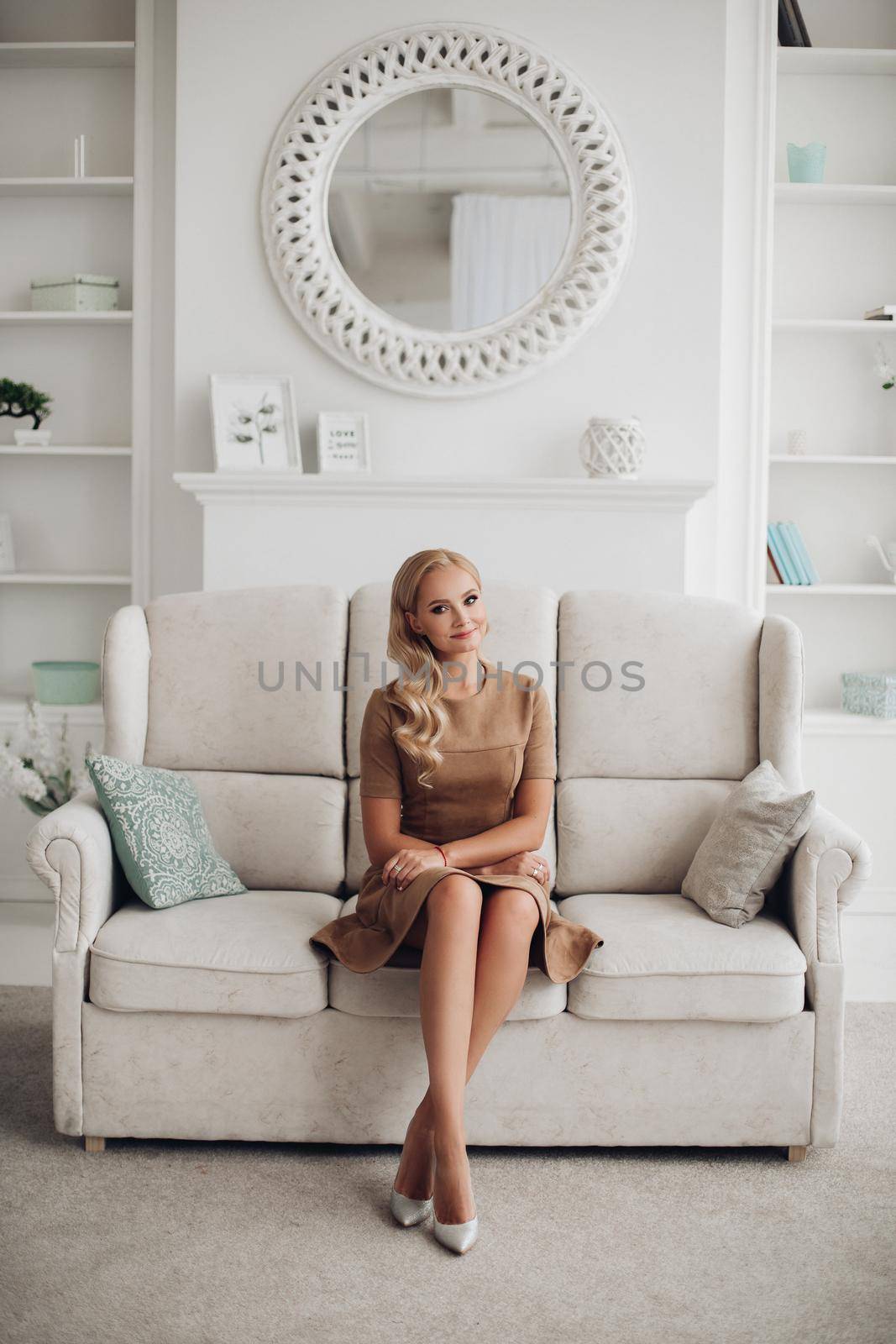 Gorgeous pretty lady sitting in luxury apartment and holding hands on knees. Beautiful young woman in elegant beige dress posing on white sofa. Happy blonde model relaxing after long working day.