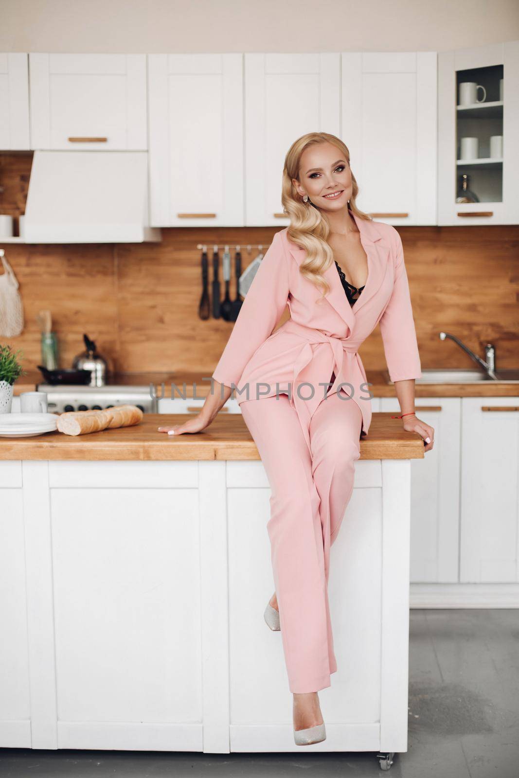 Stylish elegant attractive young blonde woman in pink suit with trousers and high heels sitting with legs crossed on kitchen counter with baguette in her hand. She is smiling at camera happily.