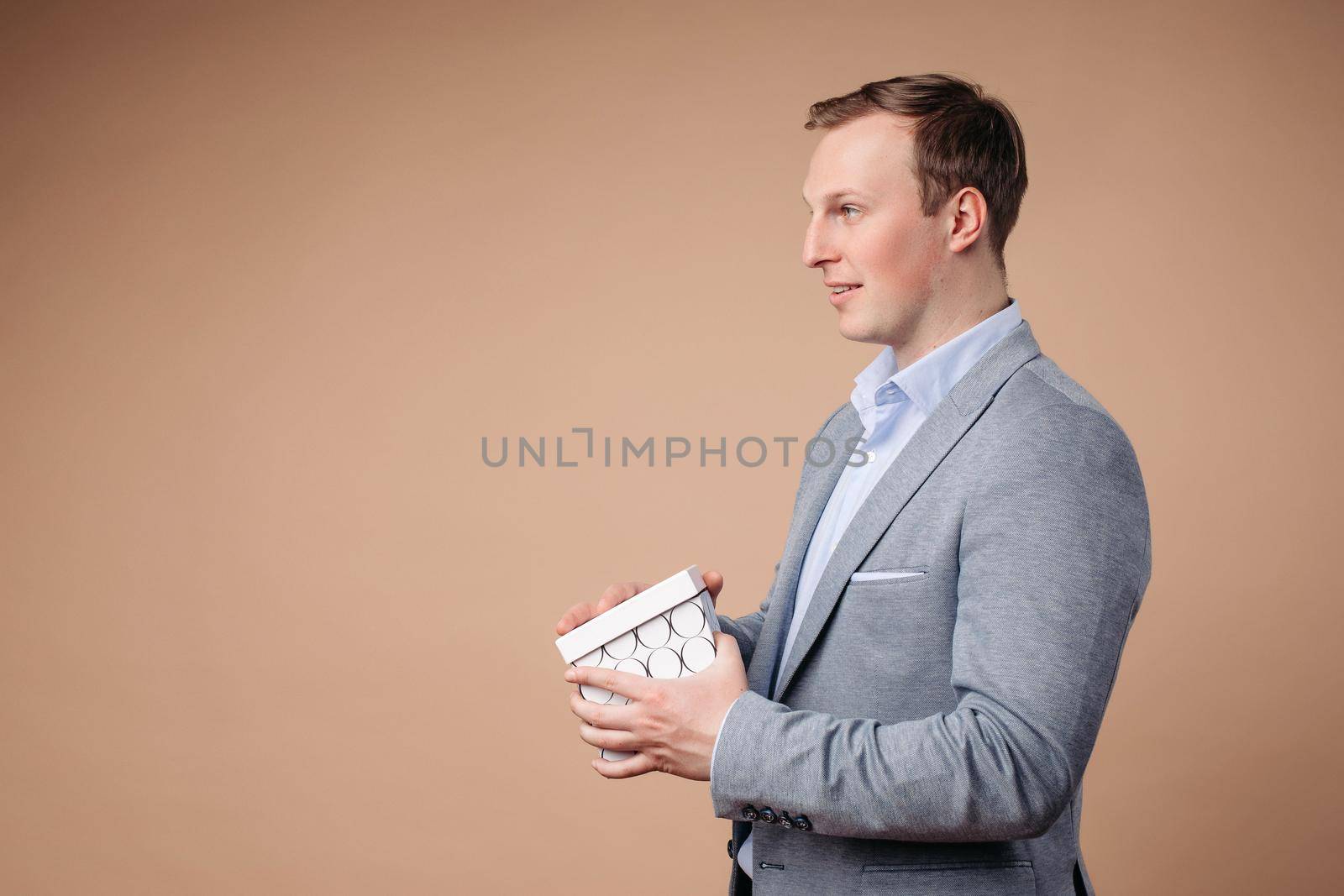 Charming caucasian male with short chestnut hair holds a white box with a gift and rejoices, picture isolated on peach background