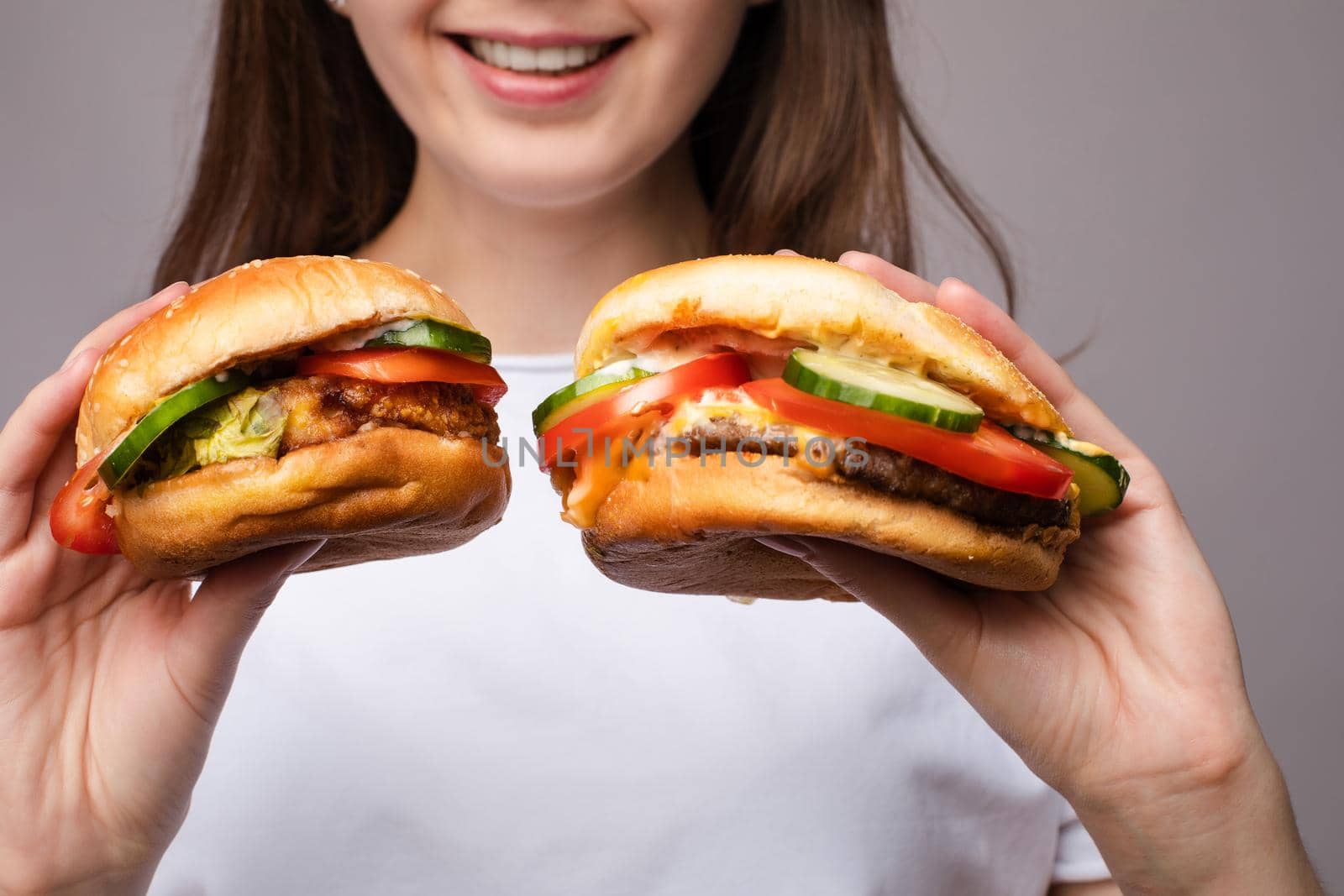 Selective focus of big tasty burgers in hands of amazed girl on grey isolated background. Shocked woman with big eyes looking at camera and eating delicious fast food. Concept of size and food.