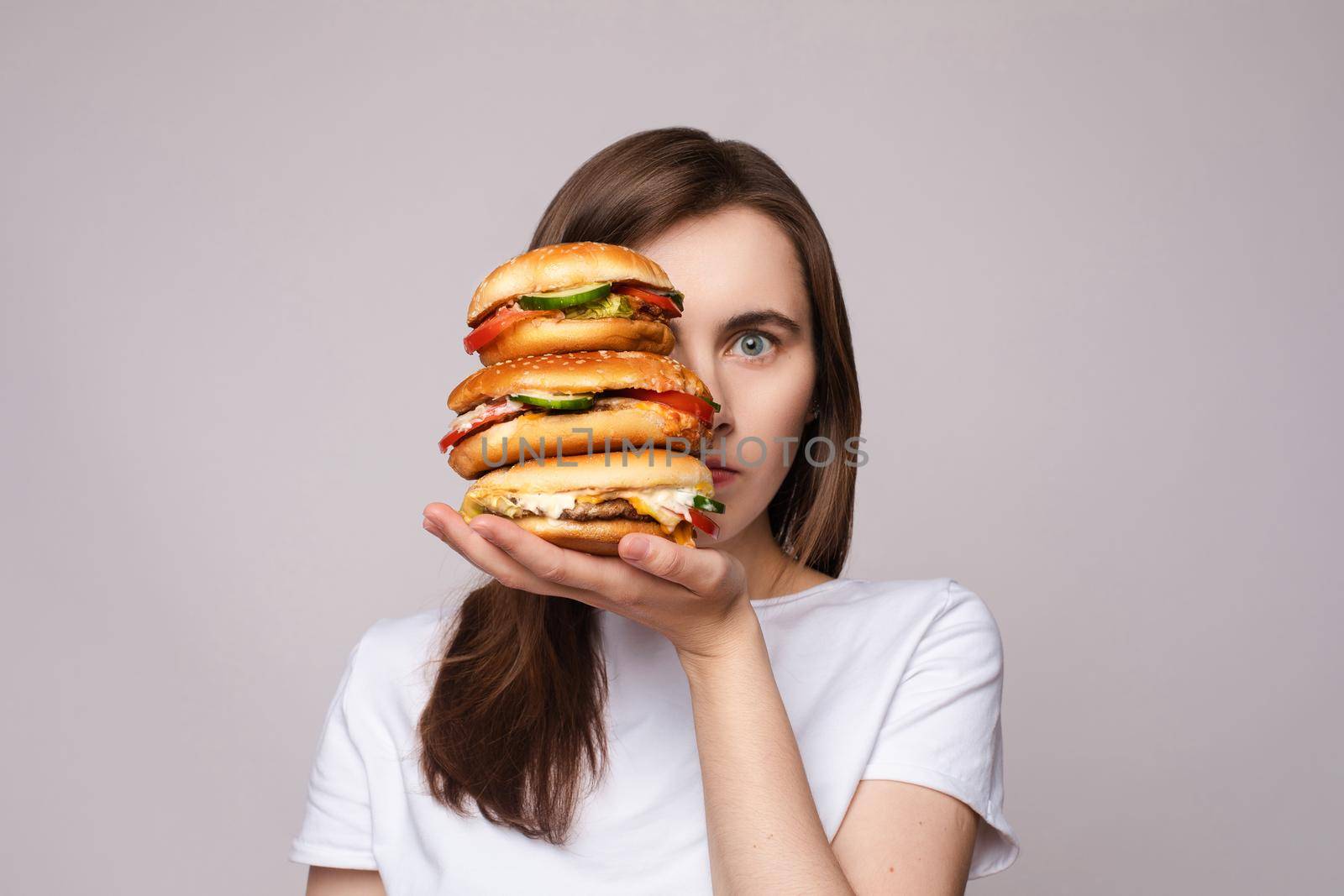 Studio portrait of young brunette woman in white t-shirt holding enormous burgers on her hand looking shocked or surprised at camera.