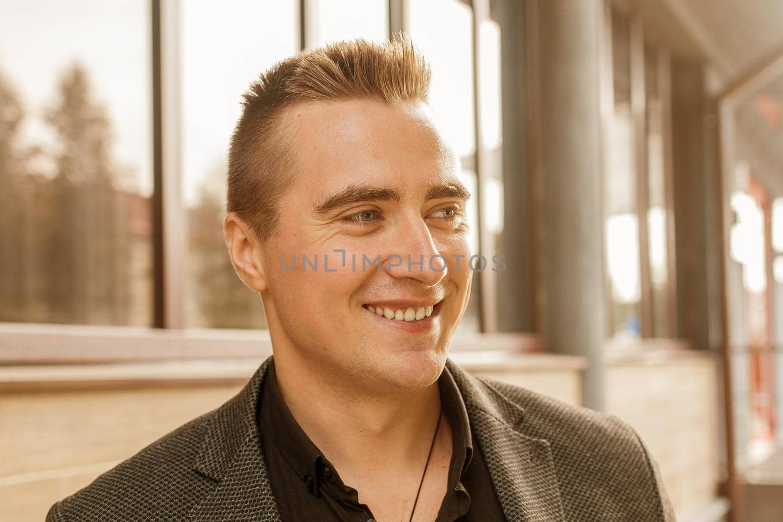 Close-up portrait of a positive smiling handsome guy of Caucasian appearance in a gray jacket and black shirt on the street outdoor.