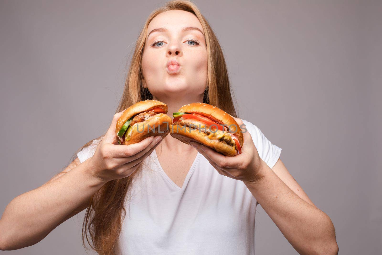 Close-up of pretty long-haired girl biting delicious burger with chicken and salad, looking at camera against pink background. American fast food concept.