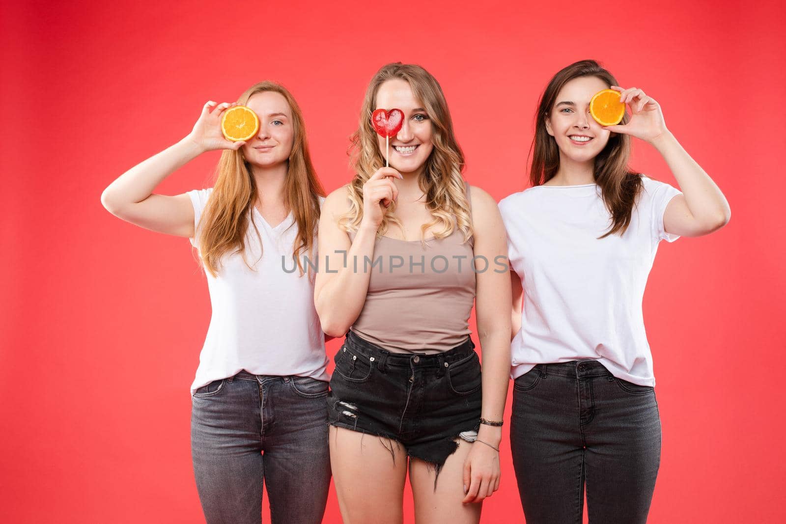Studio portrait of three cheerful pretty girlfriends holding halved oranges and one red heart-shaped lollipop. Isolate on red. Looking at camera and smile. Summer concept.