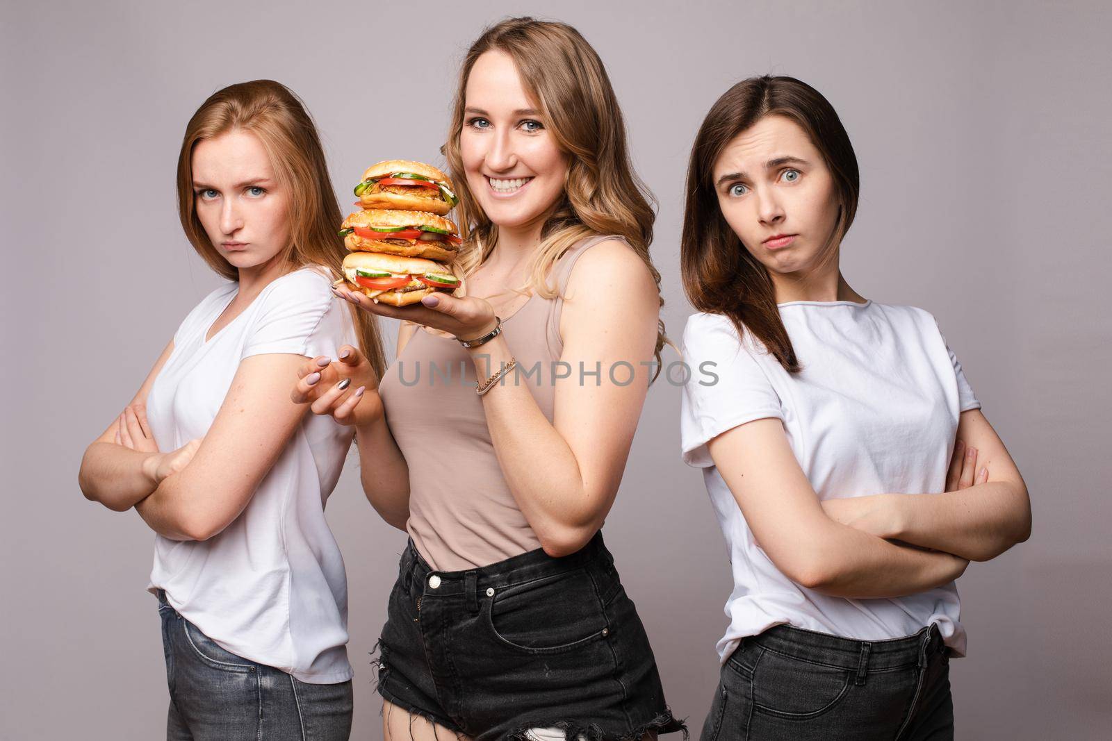 Front view of cheerful woman keeping big delicious burger and laughing while sad girls standing near and looking at fast food. Three models posing on isolated background. Concept of food and diet.