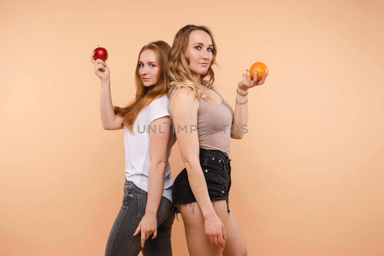 Two sporty girls looking at camera and posing with useful vegetables and fruits on isolated background. Young sisters standing back to back, keeping apple and orange and laughing.