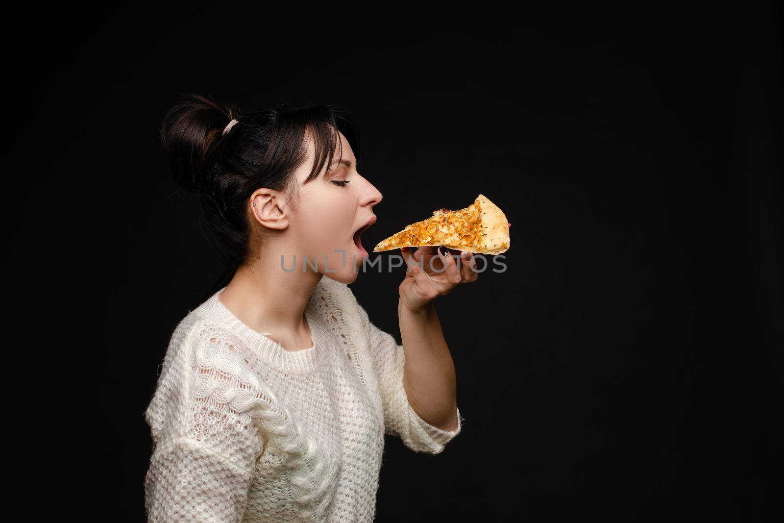 Side view portrait of brunette young girl in white sweater with tail biting delicious piece of Italian pizza. Isolate on black. Fast food concept.